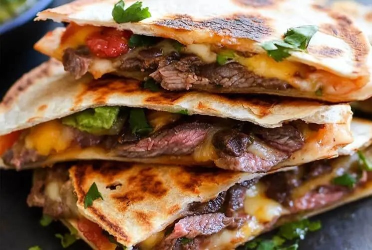 Stack of golden-brown steak quesadillas filled with melted cheese, sliced beef, avocado, and salsa, garnished with cilantro.