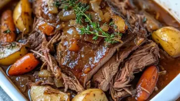 Close-up of melt in your mouth pot roast with tender beef, golden potatoes, carrots, and fresh thyme in a rich brown gravy inside a white baking dish.