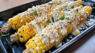 Four ears of grilled Mexican street corn topped with creamy sauce, crumbled Cotija cheese, chili powder, and fresh cilantro on a black tray.