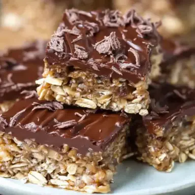 Close-up of chewy no-bake chocolate oat bars topped with melted chocolate and chocolate shavings, stacked on a white plate for a rich, homemade snack or dessert.