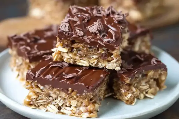 Close-up of chewy no-bake chocolate oat bars topped with melted chocolate and chocolate shavings, stacked on a white plate for a rich, homemade snack or dessert.