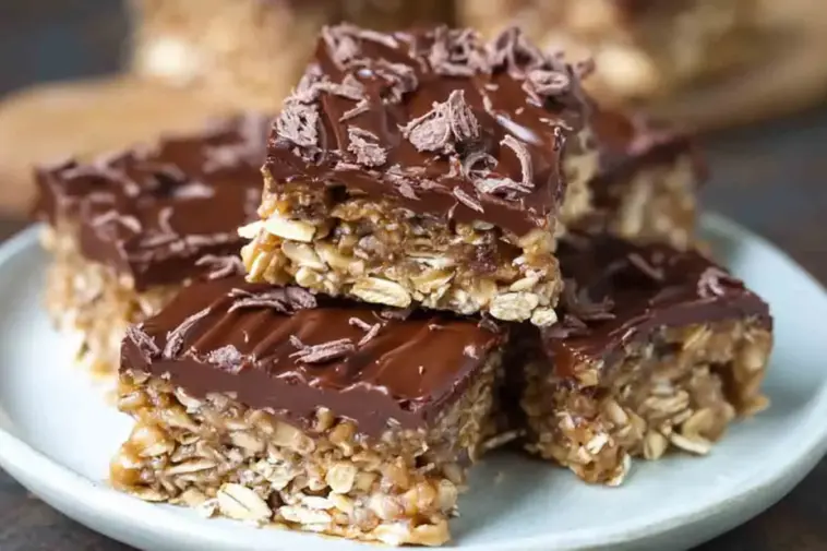Close-up of chewy no-bake chocolate oat bars topped with melted chocolate and chocolate shavings, stacked on a white plate for a rich, homemade snack or dessert.