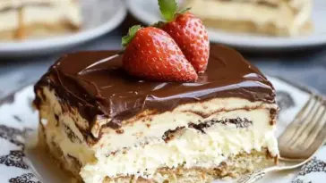 A rich slice of no-bake eclair cake with layers of vanilla pudding and graham crackers, topped with glossy chocolate frosting and fresh strawberries, served on a vintage-patterned plate.