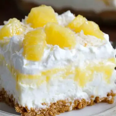 A slice of no bake pineapple dream dessert with fluffy whipped layers, graham cracker crust, and pineapple chunks on top, served on a white plate.