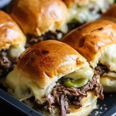 Close-up of Philly Cheesesteak Sliders with shredded beef, green peppers, and melted cheese inside golden, toasted Hawaiian rolls on a baking tray.