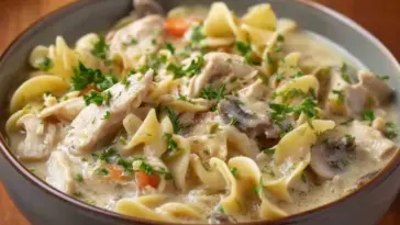 Bowl of creamy slow cooker chicken and noodles topped with parsley, featuring tender shredded chicken, egg noodles, carrots, and mushrooms in a rich broth.