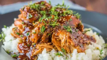 Shredded slow cooker honey garlic chicken topped with sesame seeds and chopped herbs, served over fluffy white rice in a dark bowl.