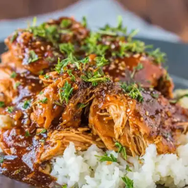 Shredded slow cooker honey garlic chicken topped with sesame seeds and chopped herbs, served over fluffy white rice in a dark bowl.