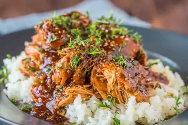 Shredded slow cooker honey garlic chicken topped with sesame seeds and chopped herbs, served over fluffy white rice in a dark bowl.
