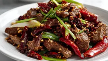 A plate of spicy cumin lamb stir fry featuring seared lamb slices, toasted cumin seeds, red chilies, and scallions, served on a white plate in a modern kitchen setting.