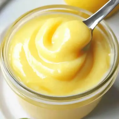 Close-up of a glass jar filled with smooth, homemade lemon curd, with a spoon dipped in and fresh lemon in the background.