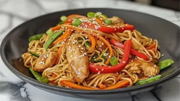 A black bowl filled with chicken lo mein on a marble counter, featuring glossy egg noodles, sliced chicken breast, red bell peppers, carrots, snow peas, and sesame seeds.