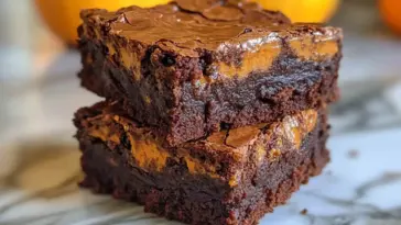 Two thick, fudgy pumpkin brownies stacked on top of each other, with a rich chocolate base and vibrant pumpkin swirl, sitting on a marble surface with small pumpkins in the blurred background.