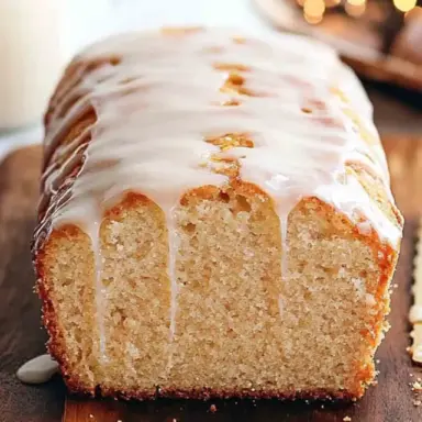 Close-up of a golden, moist eggnog bread loaf topped with a creamy glaze, sliced to reveal its soft texture, displayed on a wooden board with holiday lights in the background.