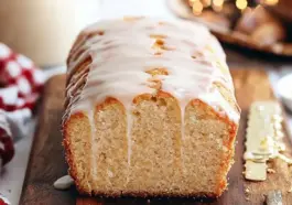 Golden eggnog bread topped with a thick glaze, sliced to show its soft, tender crumb, resting on a wooden board with festive lights and a red-checked towel in the background.
