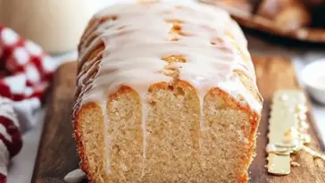 Golden eggnog bread topped with a thick glaze, sliced to show its soft, tender crumb, resting on a wooden board with festive lights and a red-checked towel in the background.