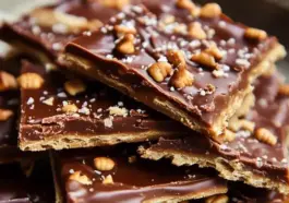 Stack of homemade Saltine Toffee pieces featuring golden caramel layers, glossy chocolate topping, and a sprinkle of chopped nuts and sea salt.