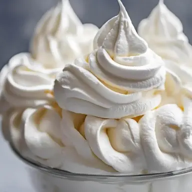 A close-up of fluffy, stabilized homemade whipped cream piped into soft peaks in a glass bowl – perfect for topping cakes, pies, and drinks.