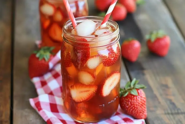 Two mason jars filled with iced Southern Strawberry Sweet Tea, loaded with sliced strawberries and ice cubes, set on a rustic wooden table with a red checkered cloth.