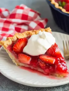Slice of Big Boy’s Fresh Strawberry Pie on a white plate, filled with glazed fresh strawberries and topped with whipped cream, with a red gingham napkin in the background.