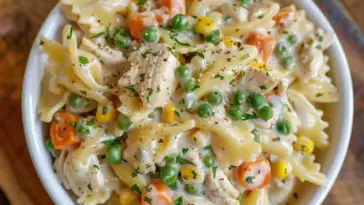 Overhead view of a bowl filled with creamy chicken pot pie pasta featuring bowtie noodles, carrots, peas, corn, and shredded chicken in a white sauce, topped with black pepper and parsley.