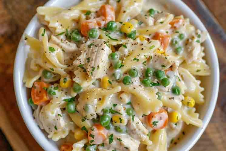Overhead view of a bowl filled with creamy chicken pot pie pasta featuring bowtie noodles, carrots, peas, corn, and shredded chicken in a white sauce, topped with black pepper and parsley.