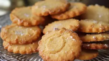 A plate of golden French butter cookies (sablés) with scalloped edges and sugar sprinkled on top, arranged on a decorative glass serving tray.