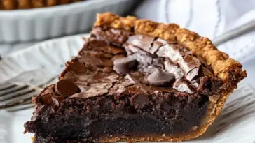 A decadent slice of gooey brownie pie with a glossy, cracked top and melted chocolate center on a white plate with chocolate chips.