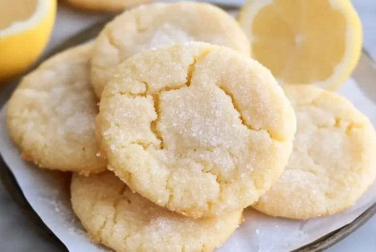 Golden lemon sugar cookies stacked on a plate, lightly coated with sugar, surrounded by fresh lemon halves.