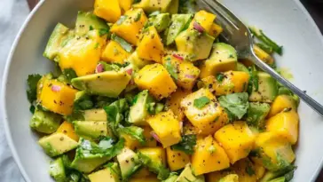 Close-up of a mango avocado salad with diced mango, avocado, red onion, and cilantro in a white bowl, served with a fork and black pepper on top