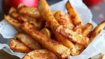 Golden brown air fryer apple fries coated in cinnamon sugar, served in a parchment-lined bowl with red apples in the background.