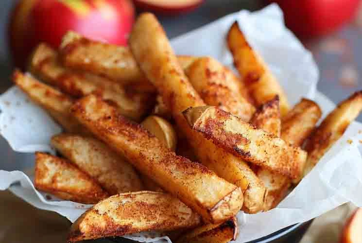 Golden brown air fryer apple fries coated in cinnamon sugar, served in a parchment-lined bowl with red apples in the background.