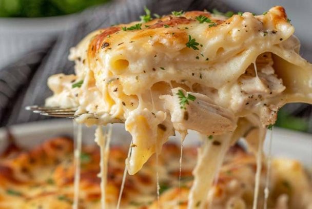A close-up of a slice of creamy white cheese chicken lasagna being lifted with a fork, showing melty mozzarella, tender shredded chicken, and golden baked layers of pasta with herbs.