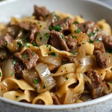 A bowl of French Onion Beef and Noodles featuring tender beef strips, caramelized onions, and wide egg noodles coated in a rich brown broth, garnished with chopped parsley.