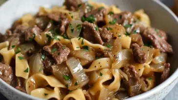 Close-up of a bowl filled with French Onion Beef and Noodles, showcasing tender beef slices, caramelized onions, and egg noodles in a rich brown sauce, topped with chopped parsley.