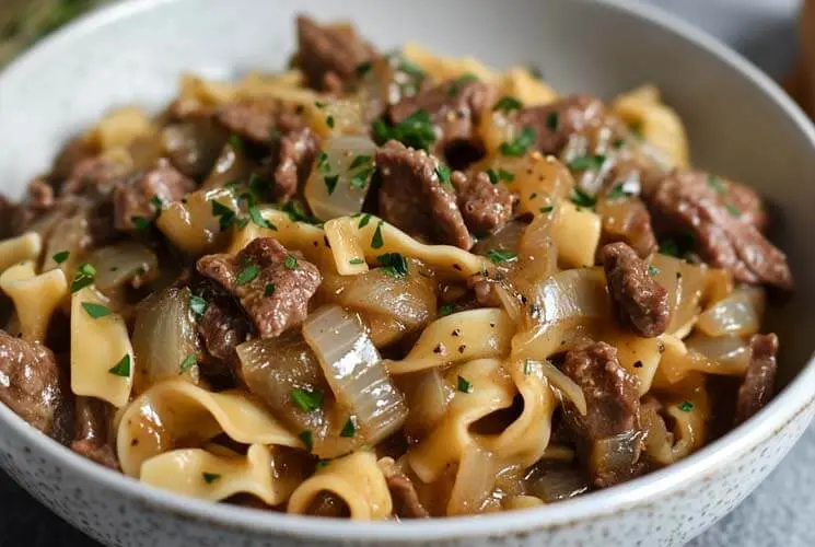 Close-up of a bowl filled with French Onion Beef and Noodles, showcasing tender beef slices, caramelized onions, and egg noodles in a rich brown sauce, topped with chopped parsley.