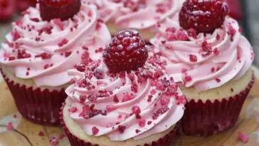 Four Raspberry Cupcakes recipe topped with piped pink frosting, crushed raspberry pieces, and whole raspberries, arranged on a wooden plate with red liners.