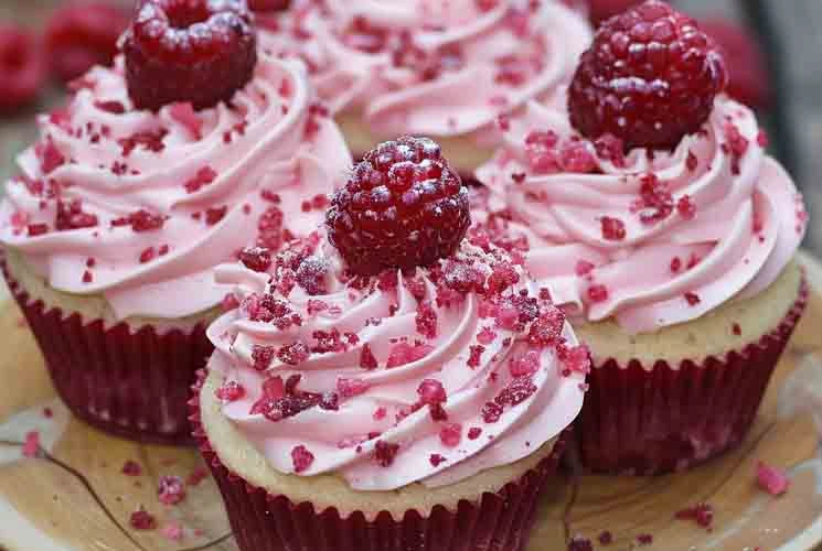 Four Raspberry Cupcakes recipe topped with piped pink frosting, crushed raspberry pieces, and whole raspberries, arranged on a wooden plate with red liners.