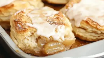 Close-up of flaky homemade biscuits filled with cinnamon apple pie filling and topped with a light glaze, baked to golden perfection in a metal pan.
