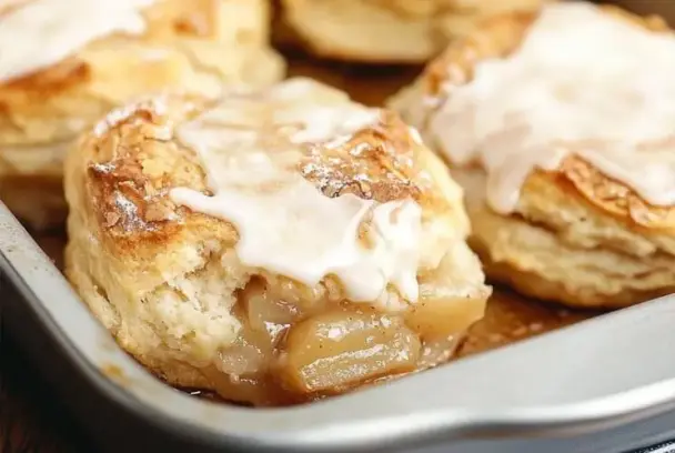 Close-up of flaky homemade biscuits filled with cinnamon apple pie filling and topped with a light glaze, baked to golden perfection in a metal pan.