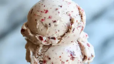 Close-up of two scoops of Dr Pepper Ice Cream with red cherry bits, showcasing a creamy texture against a blurred light background.