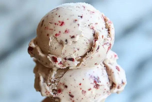 Close-up of two scoops of Dr Pepper Ice Cream with red cherry bits, showcasing a creamy texture against a blurred light background.