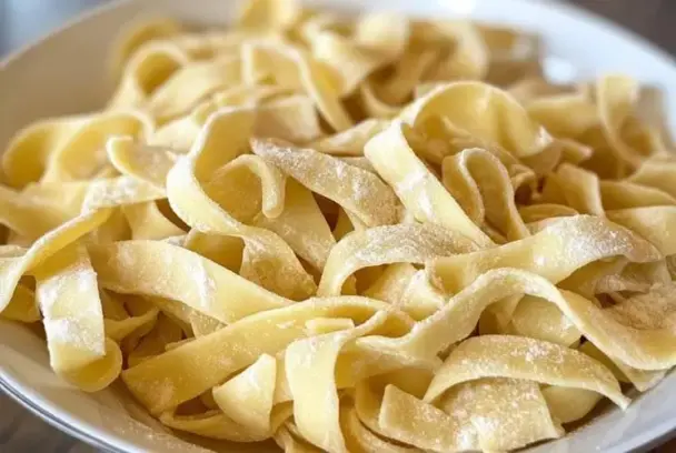 Fresh homemade egg noodles, hand-cut and lightly dusted with flour, served on a white plate, highlighting their soft, rustic texture and golden color.