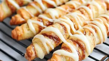 Golden-brown twisted cinnamon roll breadsticks topped with creamy white glaze, resting on a wire cooling rack.