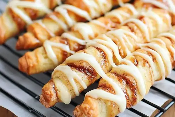 Golden-brown twisted cinnamon roll breadsticks topped with creamy white glaze, resting on a wire cooling rack.