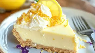 Slice of creamy no bake lemon pie with a graham cracker crust, topped with whipped cream, lemon zest, and a lemon slice, served on a white plate with a fork.