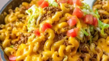 Cheesy Taco Mac and Cheese in a skillet, topped with seasoned beef, shredded lettuce, and fresh diced tomatoes, being scooped with a spoon.