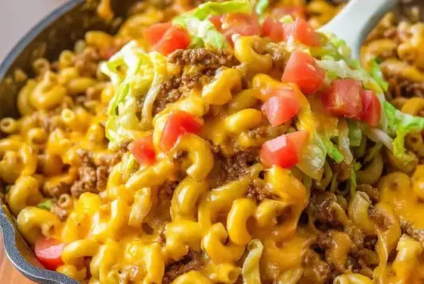 Cheesy Taco Mac and Cheese in a skillet, topped with seasoned beef, shredded lettuce, and fresh diced tomatoes, being scooped with a spoon.