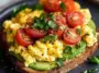 Whole grain toast layered with mashed avocado, fluffy scrambled eggs, halved cherry tomatoes, and chopped parsley on a dark plate