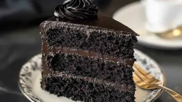 Close-up of a decadent three-layer Black Velvet Cake slice with dark chocolate frosting, served on a vintage plate with a gold fork in the background.